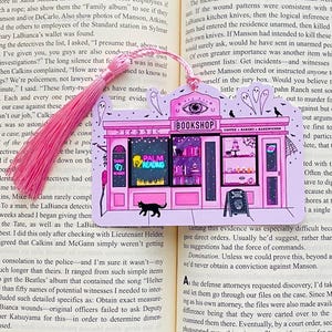 May include: Pink and black illustrated bookmark featuring a whimsical bookshop scene with the text "BOOKSHOP", "PALM READINGS", and a black cat. The bookmark has a pink tassel and is placed in an open book.