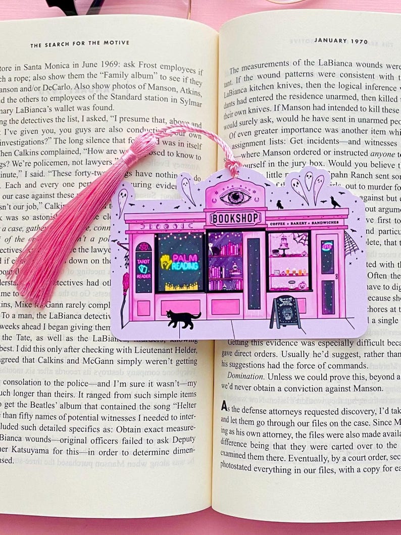 May include: Pink and black illustrated bookmark featuring a whimsical bookshop scene with the text "BOOKSHOP", "PALM READINGS", and a black cat. The bookmark has a pink tassel and is placed in an open book.