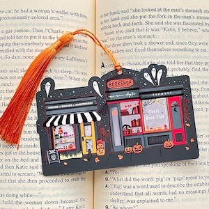 Puede incluir: Un marcap&aacute;ginas con tem&aacute;tica de Halloween que representa una panader&iacute;a y una librer&iacute;a. La panader&iacute;a tiene un toldo a rayas blancas y negras, y la librer&iacute;a est&aacute; etiquetada como "Ghost Book Club". El marcap&aacute;ginas tiene una borla naranja.