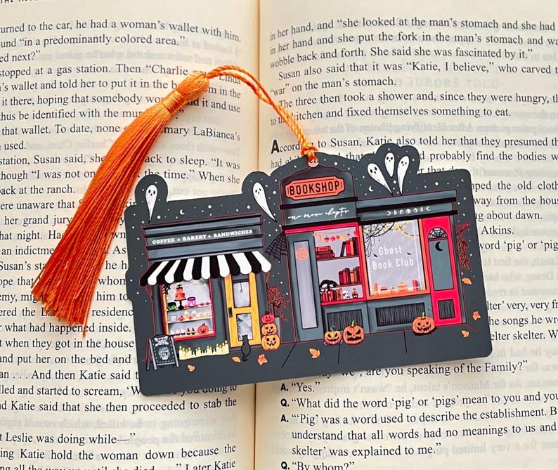 Puede incluir: Un marcap&aacute;ginas con tem&aacute;tica de Halloween que representa una panader&iacute;a y una librer&iacute;a. La panader&iacute;a tiene un toldo a rayas blancas y negras, y la librer&iacute;a est&aacute; etiquetada como "Ghost Book Club". El marcap&aacute;ginas tiene una borla naranja.