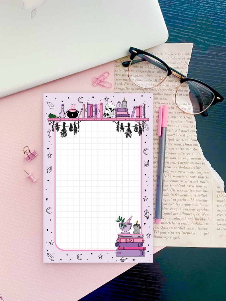 May include: A pink and white gridded notepad with a black and white illustration of a bookshelf with books, candles, a cauldron, and skulls. The border of the notepad is decorated with stars, moons, and crystals.