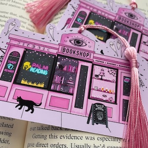May include: Pink illustrated bookmark featuring a whimsical bookshop design with the text "BOOKSHOP", "TAROT READER", and "PALM READING". The bookmark has a pink tassel and a black cat illustration. The background is a book.