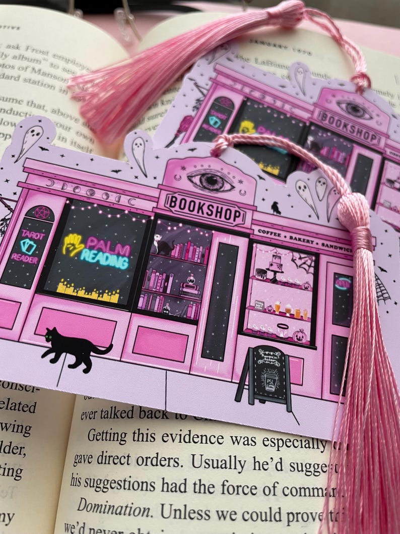 May include: Pink illustrated bookmark featuring a whimsical bookshop design with the text "BOOKSHOP", "TAROT READER", and "PALM READING". The bookmark has a pink tassel and a black cat illustration. The background is a book.