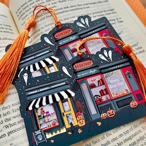 Puede incluir: Marcap&aacute;ginas azul oscuro con una escena de librer&iacute;a espeluznante con fantasmas, calabazas y el texto "BOOKSHOP" y "Ghost Book Club". El marcap&aacute;ginas tiene una borla naranja y est&aacute; sobre un libro abierto.