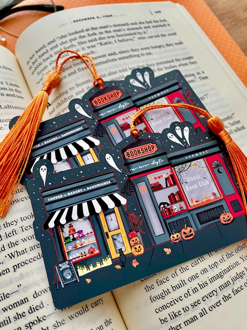 Puede incluir: Marcap&aacute;ginas azul oscuro con una escena de librer&iacute;a espeluznante con fantasmas, calabazas y el texto "BOOKSHOP" y "Ghost Book Club". El marcap&aacute;ginas tiene una borla naranja y est&aacute; sobre un libro abierto.