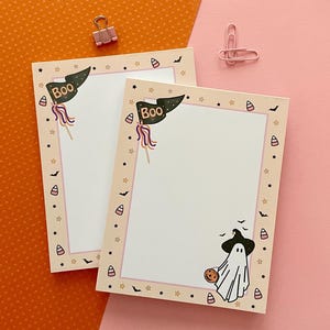 May include: Two rectangular paper notepads with a cream-colored border featuring Halloween motifs including candy corn, small black bats, and stars. The top left corner displays a dark green pennant flag with the text BOO. A ghost wearing a hat is present.
