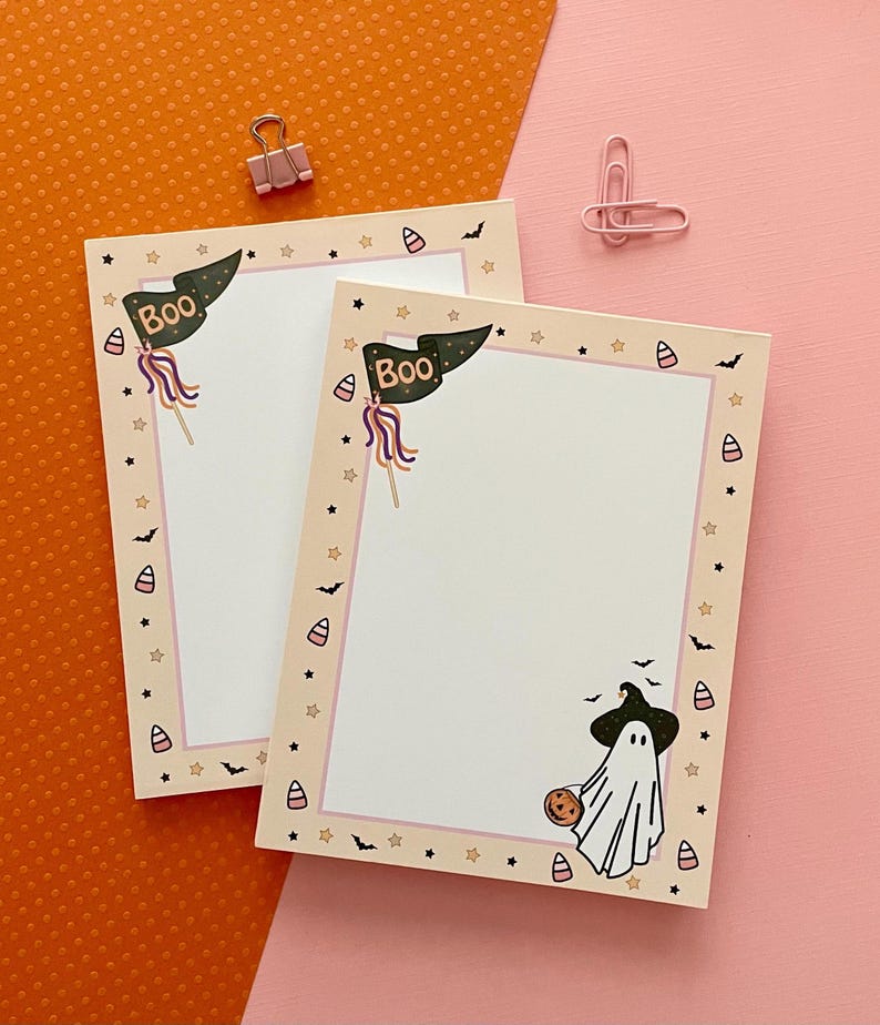 May include: Two rectangular paper notepads with a cream-colored border featuring Halloween motifs including candy corn, small black bats, and stars. The top left corner displays a dark green pennant flag with the text BOO. A ghost wearing a hat is present.