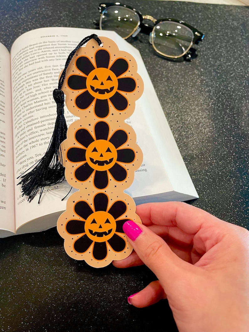 May include: A Halloween-themed bookmark featuring three orange and black pumpkin-faced flowers. The bookmark has a black tassel.