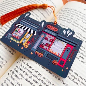 Puede incluir: Un marcap&aacute;ginas azul oscuro con un dise&ntilde;o tem&aacute;tico de Halloween que presenta una librer&iacute;a y una panader&iacute;a. El marcap&aacute;ginas tiene una borla naranja y est&aacute; colocado en un libro abierto. La librer&iacute;a tiene el texto "BOOKSHOP" y "Ghost Book Club".