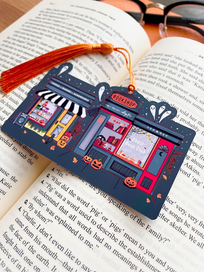 Puede incluir: Un marcap&aacute;ginas azul oscuro con un dise&ntilde;o tem&aacute;tico de Halloween que presenta una librer&iacute;a y una panader&iacute;a. El marcap&aacute;ginas tiene una borla naranja y est&aacute; colocado en un libro abierto. La librer&iacute;a tiene el texto "BOOKSHOP" y "Ghost Book Club".