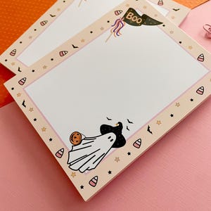 May include: A rectangular notepad with a light peach border featuring a repeating pattern of candy corn, black stars, and small bats. The top right corner displays a green pennant flag with the text Boo. A ghost wearing a hat holds a pumpkin.