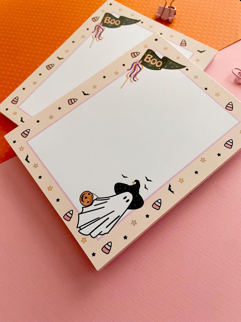 May include: A rectangular notepad with a light peach border featuring a repeating pattern of candy corn, black stars, and small bats. The top right corner displays a green pennant flag with the text Boo. A ghost wearing a hat holds a pumpkin.