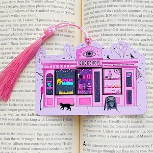 May include: Pink bookmark featuring a whimsical illustration of a bookstore with a coffee shop. The sign reads "BOOKSHOP" and "COFFEE BAKERY SANDWICHES". Includes a pink tassel and the words "TAROT READER" and "PALM READING".