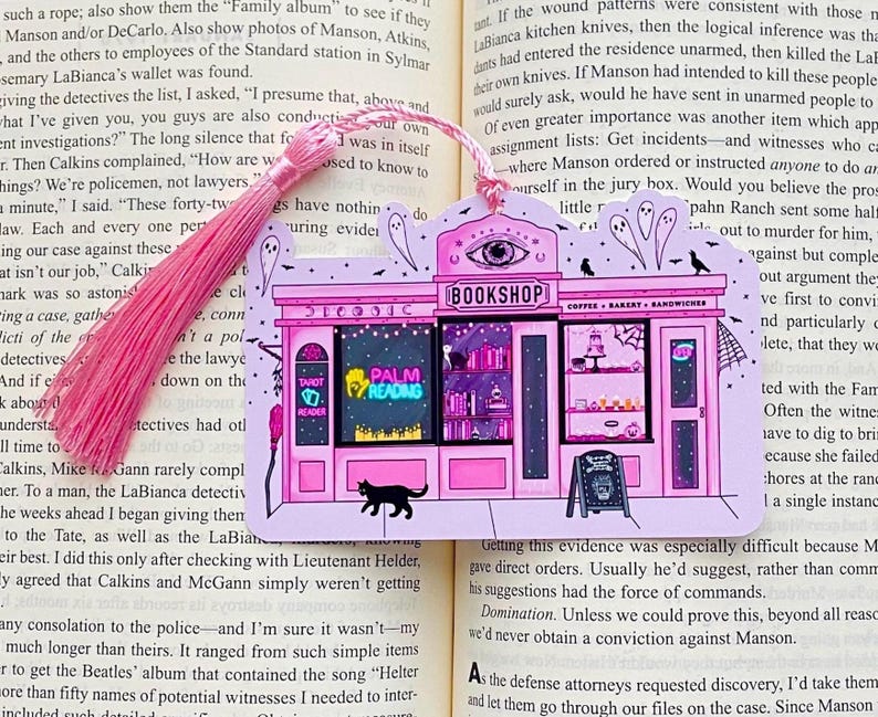 May include: Pink bookmark featuring a whimsical illustration of a bookstore with a coffee shop. The sign reads "BOOKSHOP" and "COFFEE BAKERY SANDWICHES". Includes a pink tassel and the words "TAROT READER" and "PALM READING".