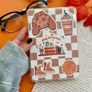 May include: A clear phone case with a brown and white checkered pattern, adorned with stickers. Stickers include a sweater, coffee drink, boots, pumpkin, and the text "Cozy Bookclub". The case is held in a person's hand.