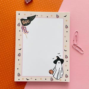 May include: A rectangular notepad with a cream-colored border featuring Halloween-themed illustrations including a black flag with the text Boo, a ghost wearing a witch hat holding a pumpkin, candy corn, bats, and stars, accompanied by two pink paper clips.