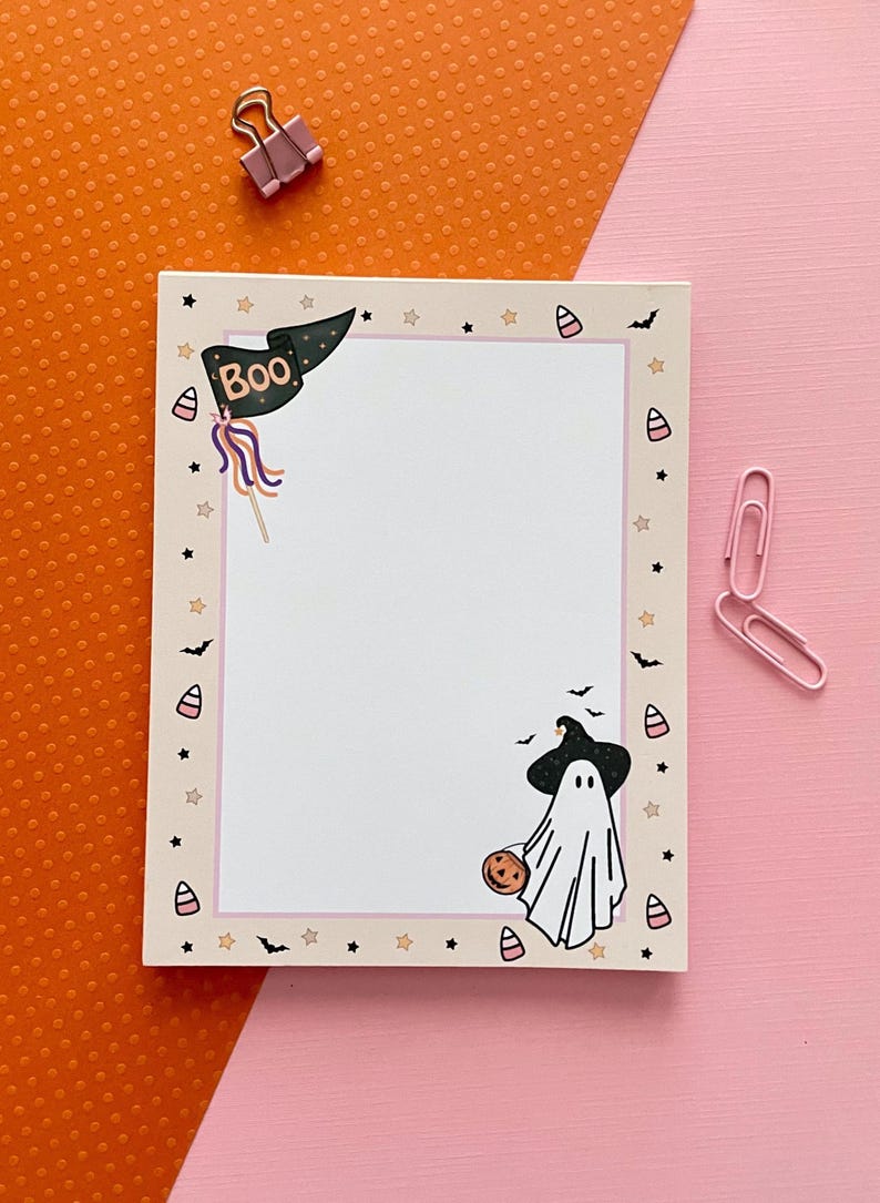 May include: A rectangular notepad with a cream-colored border featuring Halloween-themed illustrations including a black flag with the text Boo, a ghost wearing a witch hat holding a pumpkin, candy corn, bats, and stars, accompanied by two pink paper clips.