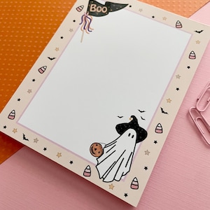 May include: A rectangular notepad with a white center and a light beige border decorated with small black bats, stars, and candy corn icons. The top features a black pennant flag with the text Boo, and the bottom shows a ghost wearing a witch hat.