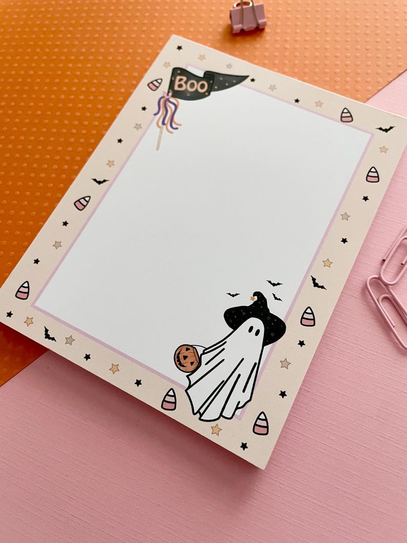 May include: A rectangular notepad with a white center and a light beige border decorated with small black bats, stars, and candy corn icons. The top features a black pennant flag with the text Boo, and the bottom shows a ghost wearing a witch hat.