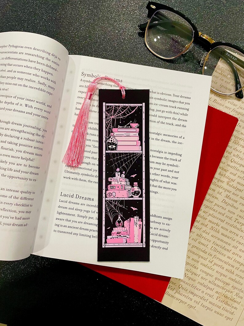 May include: A black bookmark featuring a pink and white illustration of a bookshelf with books, candles, and potion bottles. The bookshelf is decorated with a spiderweb and bats. The bookmark has a pink tassel.