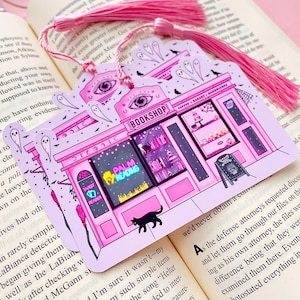 May include: Pink and purple illustrated bookmark featuring a whimsical bookshop design with the text "BOOKSHOP", "PALM READING", and "TAROT READER". Includes a black cat, ghosts, and a pink tassel.
