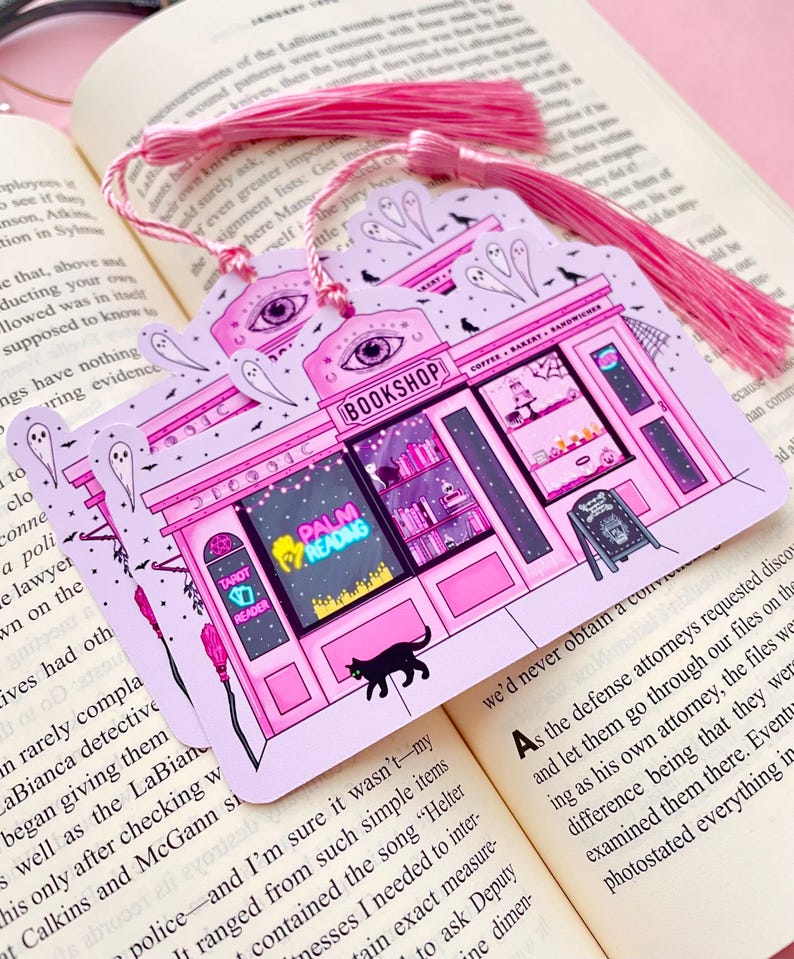 May include: Pink and purple illustrated bookmark featuring a whimsical bookshop design with the text "BOOKSHOP", "PALM READING", and "TAROT READER". Includes a black cat, ghosts, and a pink tassel.