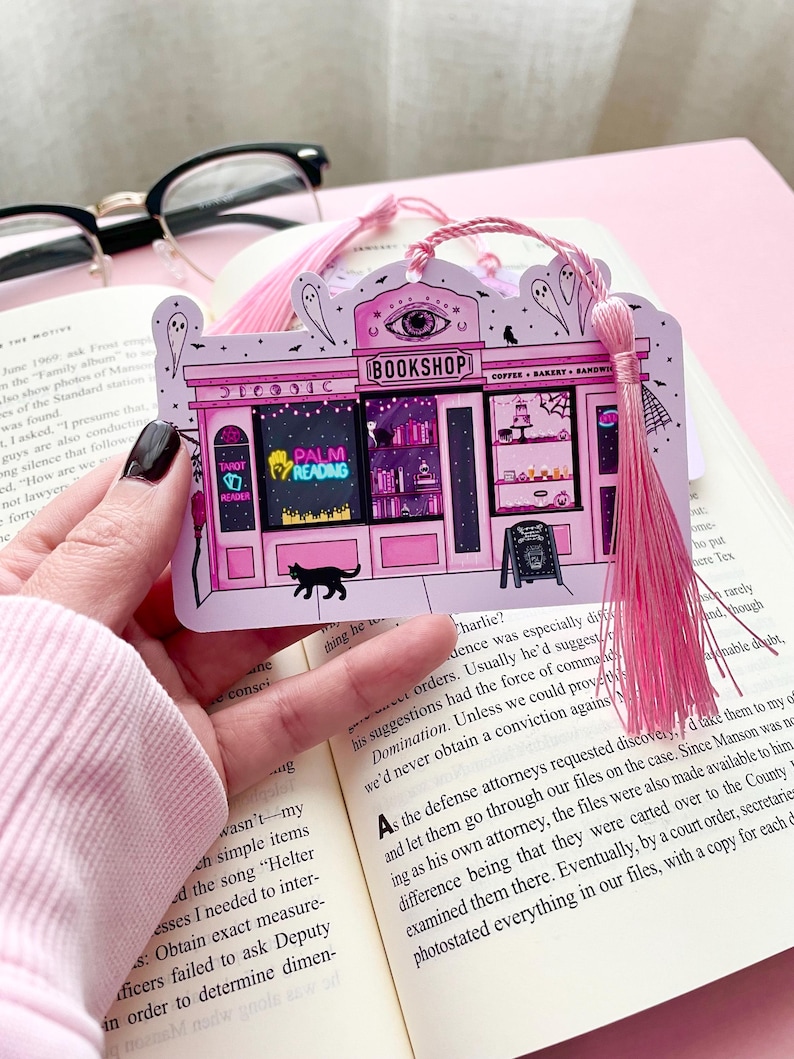 May include: Pink bookmark featuring a whimsical illustration of a bookshop with a black cat, ghosts, and the text "BOOKSHOP". Includes a pink tassel. The bookmark is held over an open book.