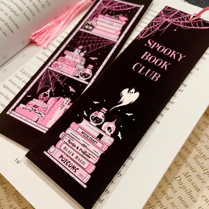 May include: Two black bookmarks with pink accents and a pink tassel. The bookmarks feature a spooky book club theme with a stack of books, a potion bottle, bats, and a ghost. The text on the bookmarks reads "Spooky Book Club".