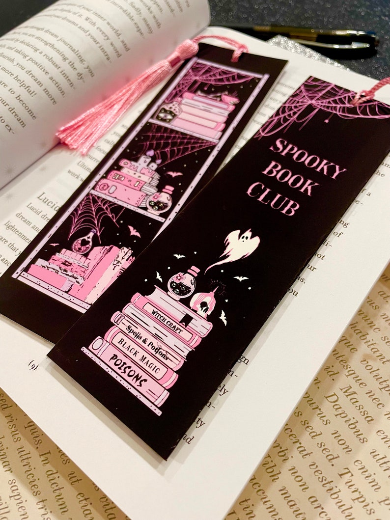 May include: Two black bookmarks with pink accents and a pink tassel. The bookmarks feature a spooky book club theme with a stack of books, a potion bottle, bats, and a ghost. The text on the bookmarks reads "Spooky Book Club".