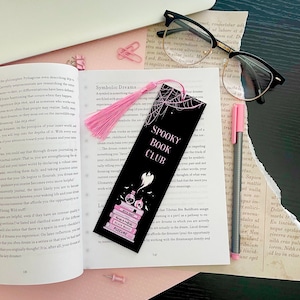 May include: A black bookmark with a pink tassel features a spooky book club theme. The bookmark has a black and white illustration of a ghost, a stack of books, and a potion bottle. The text "Spooky Book Club" is written in pink letters on the bookmark.