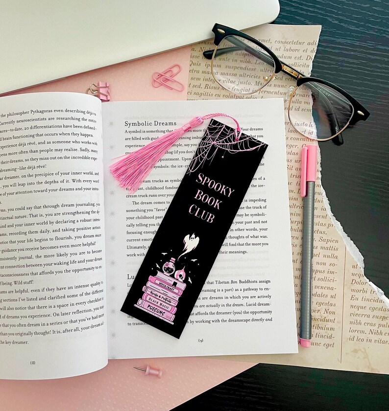 May include: A black bookmark with a pink tassel features a spooky book club theme. The bookmark has a black and white illustration of a ghost, a stack of books, and a potion bottle. The text "Spooky Book Club" is written in pink letters on the bookmark.
