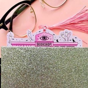 May include: A pink and white bookmark with a tassel, featuring a spooky "BOOKSHOP" design with an eye symbol, ghosts, and birds. The bookmark is inserted into a glittery silver notebook, with eyeglasses in the background.