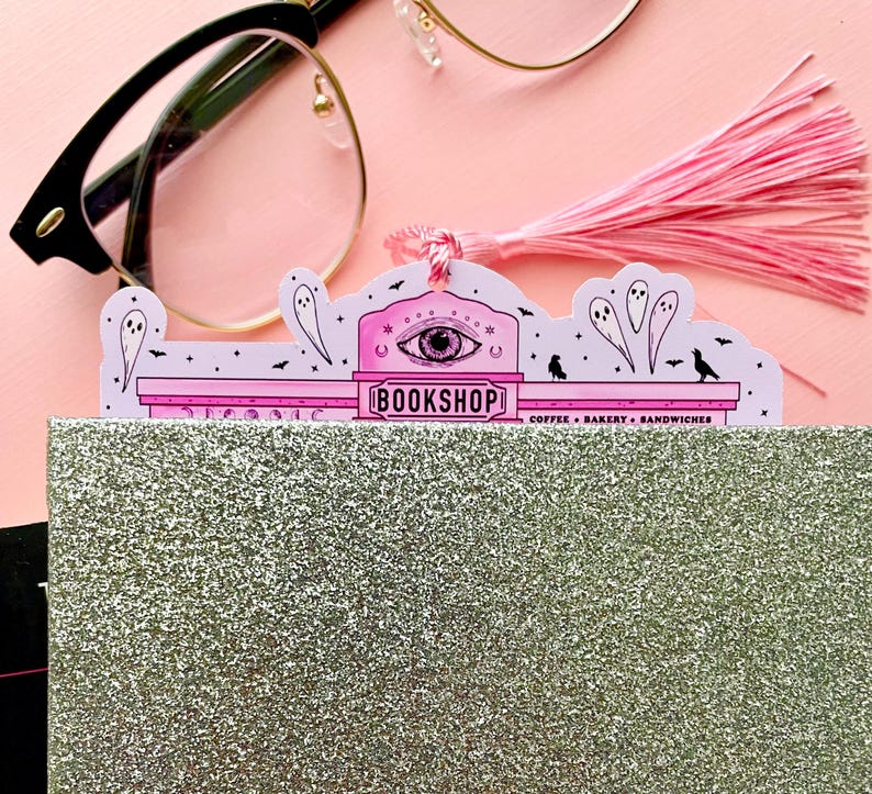 May include: A pink and white bookmark with a tassel, featuring a spooky "BOOKSHOP" design with an eye symbol, ghosts, and birds. The bookmark is inserted into a glittery silver notebook, with eyeglasses in the background.