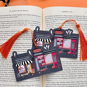 Puede incluir: Tres marcap&aacute;ginas con tem&aacute;tica de Halloween con borlas naranjas descansan sobre un libro abierto. Cada marcap&aacute;ginas presenta una fachada de tienda con una librer&iacute;a y un caf&eacute;, fantasmas y calabazas. Los textos son "Bookshop" y "Ghost Book Club".