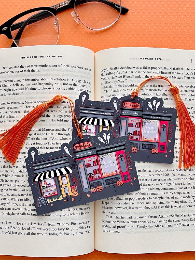 Puede incluir: Tres marcap&aacute;ginas con tem&aacute;tica de Halloween con borlas naranjas descansan sobre un libro abierto. Cada marcap&aacute;ginas presenta una fachada de tienda con una librer&iacute;a y un caf&eacute;, fantasmas y calabazas. Los textos son "Bookshop" y "Ghost Book Club".