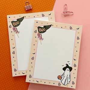 May include: Two rectangular paper notepads featuring a cream-colored border with a pattern of small stars, bats, and candy corn. Each notepad displays a dark green pennant flag with the text BOO and a white ghost wearing a black witch hat.