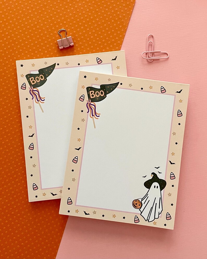 May include: Two rectangular paper notepads featuring a cream-colored border with a pattern of small stars, bats, and candy corn. Each notepad displays a dark green pennant flag with the text BOO and a white ghost wearing a black witch hat.