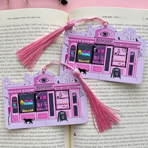 May include: Two pink and white illustrated bookmarks with pink tassels. Each bookmark depicts a whimsical bookshop with the text "BOOKSHOP", "PALM READING", and "COFFEE BAKERY SANDWICHES". Black cat and ghost illustrations.