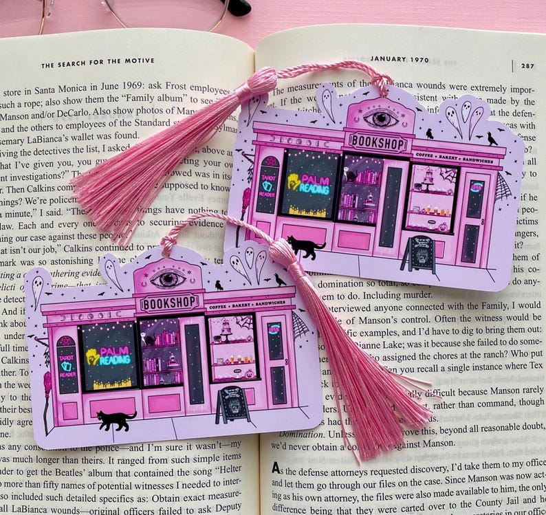 May include: Two pink and white illustrated bookmarks with pink tassels. Each bookmark depicts a whimsical bookshop with the text "BOOKSHOP", "PALM READING", and "COFFEE BAKERY SANDWICHES". Black cat and ghost illustrations.