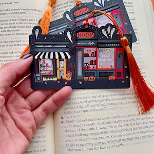 Puede incluir: Marcap&aacute;ginas negro con una ilustraci&oacute;n de librer&iacute;a, con el texto "BOOKSHOP" y "Ghost Book Club". El dise&ntilde;o incluye fantasmas, calabazas y una cafeter&iacute;a. Una borla naranja est&aacute; adjunta. Ideal para los amantes de los libros.