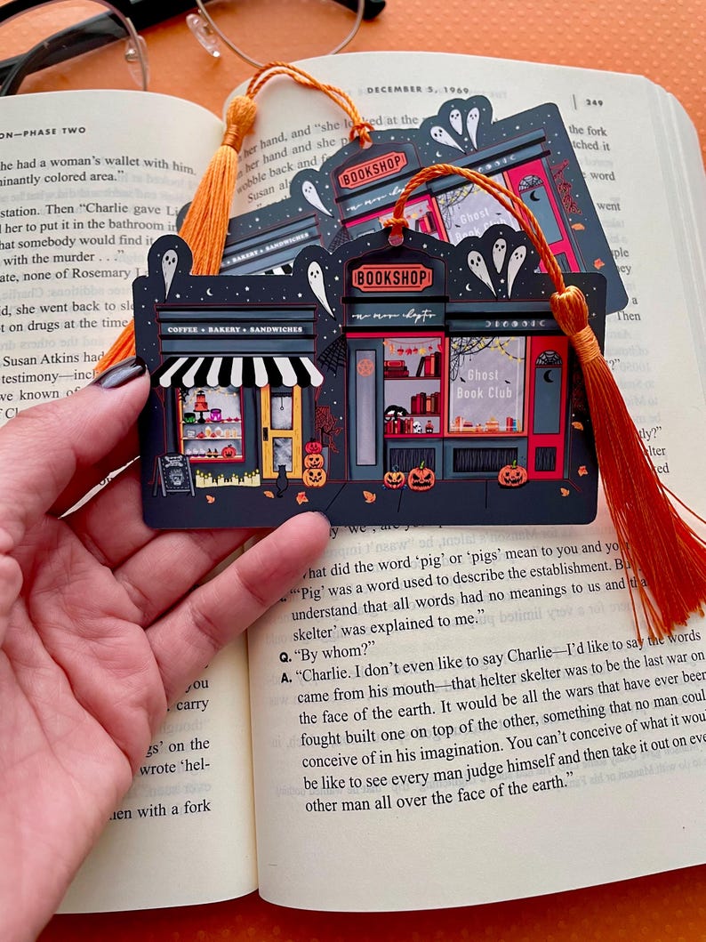 Puede incluir: Marcap&aacute;ginas negro con una ilustraci&oacute;n de librer&iacute;a, con el texto "BOOKSHOP" y "Ghost Book Club". El dise&ntilde;o incluye fantasmas, calabazas y una cafeter&iacute;a. Una borla naranja est&aacute; adjunta. Ideal para los amantes de los libros.