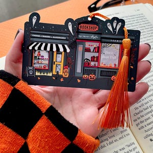 Puede incluir: Un marcap&aacute;ginas negro con tem&aacute;tica de Halloween que representa una librer&iacute;a y un caf&eacute;. El marcap&aacute;ginas tiene una borla naranja y contiene el texto "BOOKSHOP", "Ghost Book Club" y "COFFEE &bull; BAKERY &bull; SANDWICHES". Las calabazas y los fantasmas decoran la escena.