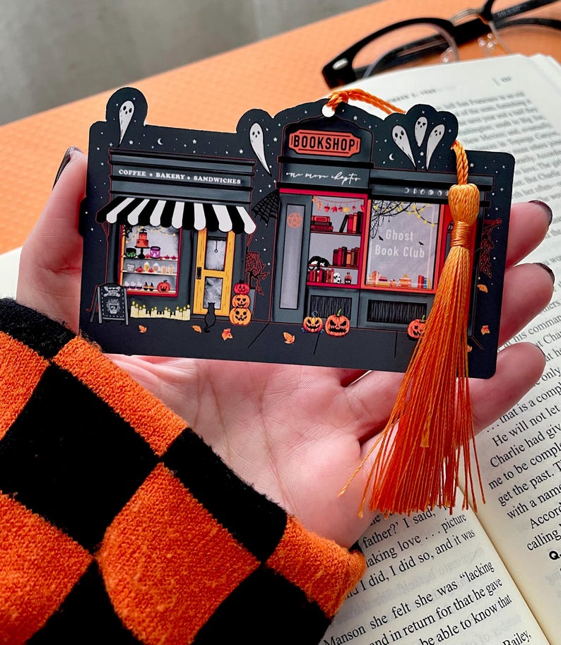 Puede incluir: Un marcap&aacute;ginas negro con tem&aacute;tica de Halloween que representa una librer&iacute;a y un caf&eacute;. El marcap&aacute;ginas tiene una borla naranja y contiene el texto "BOOKSHOP", "Ghost Book Club" y "COFFEE &bull; BAKERY &bull; SANDWICHES". Las calabazas y los fantasmas decoran la escena.