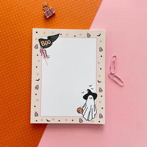 May include: A rectangular notepad featuring a cream-colored border decorated with small stars, bats, and candy corn. The top left corner displays a black pennant flag with the text BOO, while the bottom right corner shows a ghost wearing a witch hat.