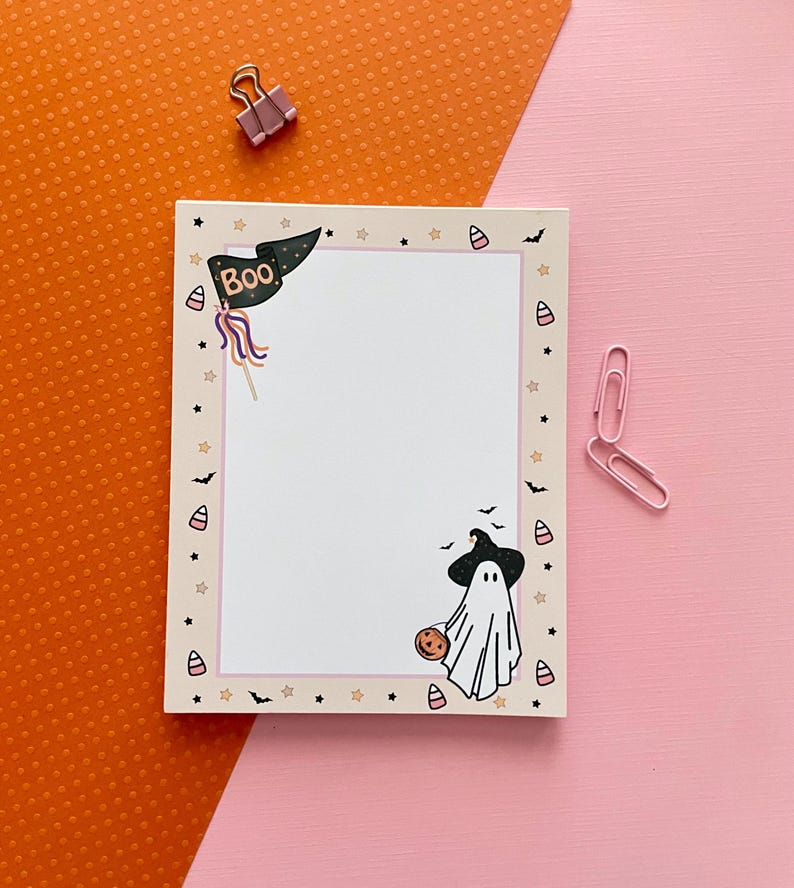 May include: A rectangular notepad featuring a cream-colored border decorated with small stars, bats, and candy corn. The top left corner displays a black pennant flag with the text BOO, while the bottom right corner shows a ghost wearing a witch hat.