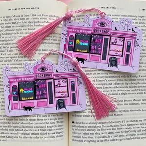 May include: Two pink bookmarks with pink tassels. Each bookmark features a pink illustration of a whimsical bookshop with the text "BOOKSHOP" and "PALM READING". A black cat and ghosts are also depicted.