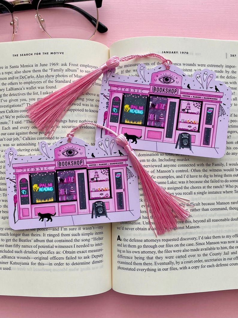 May include: Two pink bookmarks with pink tassels. Each bookmark features a pink illustration of a whimsical bookshop with the text "BOOKSHOP" and "PALM READING". A black cat and ghosts are also depicted.