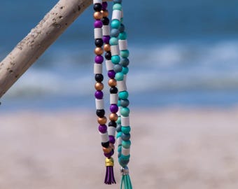 May include: Two beaded necklaces with tassels hang from a piece of driftwood on a sandy beach. One necklace features purple, black, copper, and white beads. The other has teal, gray, and white beads. The ocean is in the background.