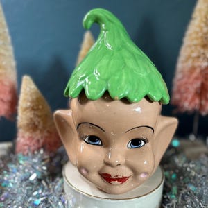 May include: A ceramic elf head figurine with a green leaf hat. The elf has a rosy complexion, blue eyes, and a painted smile.