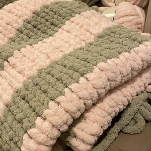 May include: A handmade, chunky knit blanket in a checkerboard pattern of soft pink and muted green. The blanket is made with thick, plush yarn, and several skeins of yarn are visible in the background.